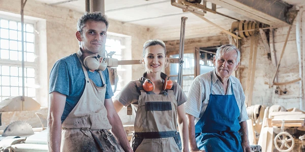 Drei Handwerker stehen in einer Werkstatt, umgeben von Werkzeugen und Maschinen. Der Mann links trägt Ohrenschützer und hat eine Schürze an, die Frau in der Mitte trägt ebenfalls Ohrenschützer und eine Arbeitskleidung, während der ältere Mann rechts eine blaue Schürze trägt. Alle lächeln in die Kamera.