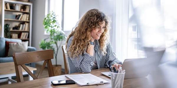 Eine Frau mit lockigem Haar sitzt an einem Holztisch und arbeitet an einem Laptop. Sie trägt eine gestreifte Bluse und lächelt, während sie den Bildschirm betrachtet. Neben ihr liegen Notizen und ein Smartphone. Im Hintergrund sind ein Regal mit Büchern und eine Zimmerpflanze zu sehen.