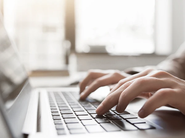 Eine Nahaufnahme von Händen, die auf einer Laptop-Tastatur tippen, während im Hintergrund weiches, natürliches Licht durch ein Fenster strömt.
