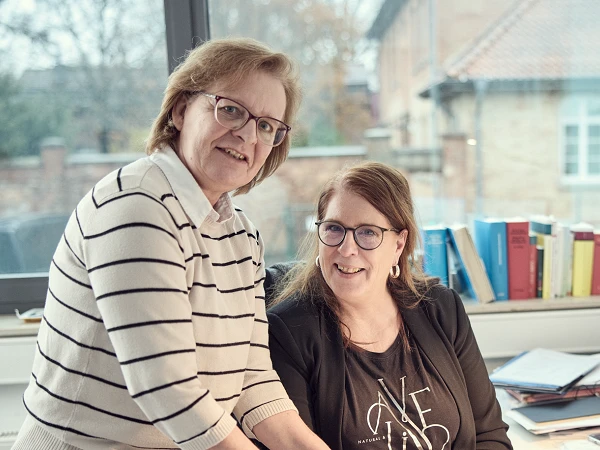Zwei Frauen stehen und sitzen an einem Schreibtisch in einem Büro, während sie in ein Dokument schauen. Im Hintergrund sind Bücherregale und eine große Fensterfront sichtbar.