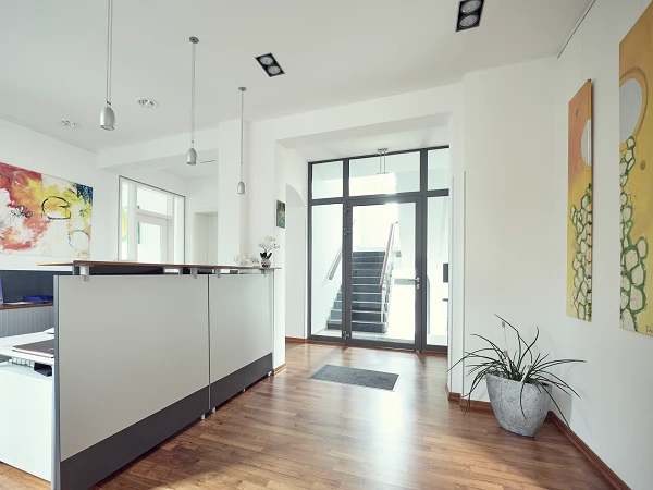 Der Empfangsbereich eines modernen Büros mit einem hellen, offenen Raumdesign. Eine Empfangstheke steht im Vordergrund, und im Hintergrund sind zwei farbenfrohe Gemälde sowie eine Treppe sichtbar, die zu einem weiteren Bereich führt. Eine Pflanze steht in der Ecke, und es gibt große Fenster, die viel Tageslicht hereinlassen.
