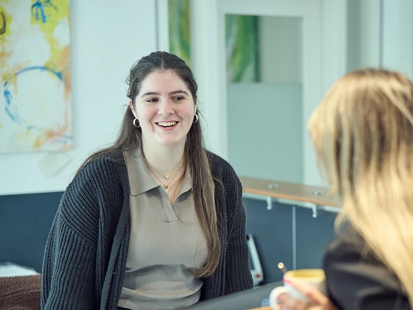 Eine junge Frau mit langen, braunen Haaren und einem lächelnden Gesicht steht an einem Empfangstresen und spricht freundlich mit einer anderen Person, die mit dem Rücken zur Kamera steht. Im Hintergrund sind einige Dekorationen und ein paar Informationsmaterialien sichtbar.