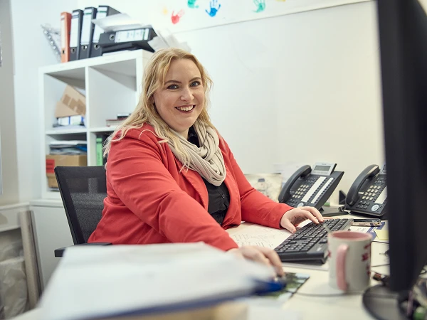 Eine lächelnde Frau in einem roten Blazer sitzt an einem Schreibtisch, tippt auf einer Tastatur und schaut in die Kamera. Im Hintergrund sind mehrere Telefone und Gegenstände auf dem Schreibtisch sichtbar.