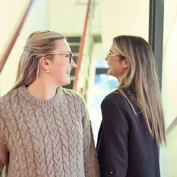 Zwei Frauen stehen in einem hellen Raum und blicken freundlich zueinander. Eine Frau trägt einen großen, strickartigen Pullover, während die andere eine dunkle Jacke hat. Im Hintergrund sind Treppen und eine moderne Architektur sichtbar.