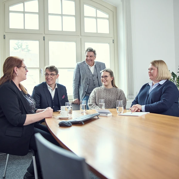 Eine Gruppe von fünf Personen sitzt an einem Konferenztisch in einem hellen Raum mit großen Fenstern. Vier Personen sind am Tisch platziert und in ein Gespräch vertieft, während eine Person hinter ihnen steht und aufmerksam lauscht. Auf dem Tisch stehen Gläser mit Wasser und Unterlagen. Pflanzen sind im Hintergrund zu sehen.