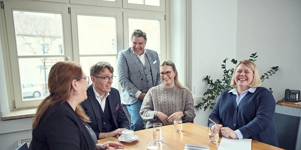 Eine Gruppe von fünf Personen sitzt an einem Tisch in einem hellen Raum, während sie sich unterhalten und Lächeln austauschen. Auf dem Tisch stehen Kaffeetassen und Gläser mit Wasser. Jeder ist in formeller Kleidung, und im Hintergrund sind Fenster und eine Pflanze zu sehen.