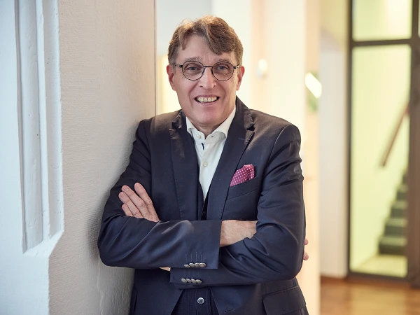 Ein Mann mit Brille und grauen Haaren leans an einer Wand in einem modernen Innenraum und lächelt. Er trägt einen eleganten Anzug mit einer Weste und hat ein rotes Taschentuch in der Brusttasche. Im Hintergrund sind Treppen und ein heller Flur sichtbar.