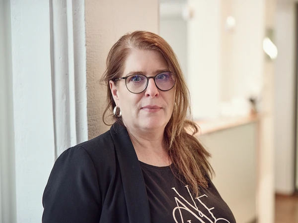 Eine Frau mit langen, braunen Haaren und Brille steht an einer Wand in einem modernen Innenraum und schaut direkt in die Kamera. Sie trägt ein schwarzes Oberteil mit einem weißen Schriftzug und eine schwarze Jacke. Im Hintergrund sind unscharfe Möbel und Lichtquellen zu erkennen.