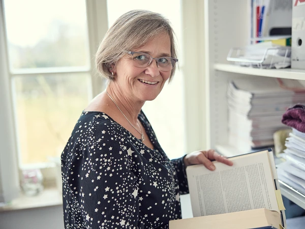 Eine lächelnde Frau mit Brille steht neben einem Regal und hält ein aufgeschlagenes Buch in der Hand, während sie nach einem weiteren Buch greift. Im Hintergrund sind Ordner und Papierstapel zu sehen.