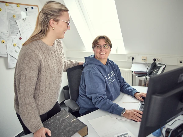 Zwei Frauen in einem Büro kommunizieren, während eine von ihnen am Computer sitzt und die andere hinter ihr steht und lächelt. Die sitzende Frau trägt einen blauen Kapuzenpullover, während die stehende Frau einen beigen Pullover trägt und eine Brille auf hat. Ein Schreibtisch mit Dokumenten und einem Computer ist im Vordergrund sichtbar.