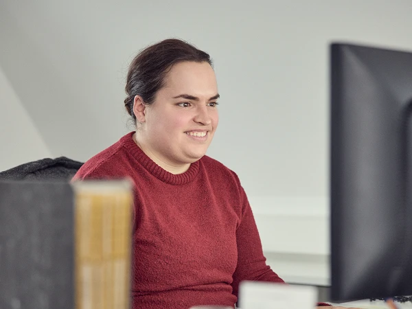 Eine Person mit langen, dunklen Haaren und einem roten Pullover sitzt an einem Schreibtisch und arbeitet konzentriert an einem Computer. Auf dem Tisch befinden sich verschiedene Büromaterialien und ein leeres Glas.