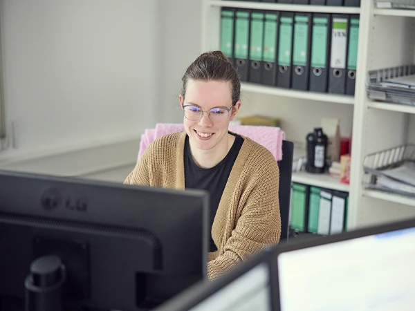 Eine junge Frau mit Brille sitzt an einem Schreibtisch und lächelt, während sie auf einen Computerbildschirm schaut. Im Hintergrund sind Aktenordner und Büromaterial zu sehen.