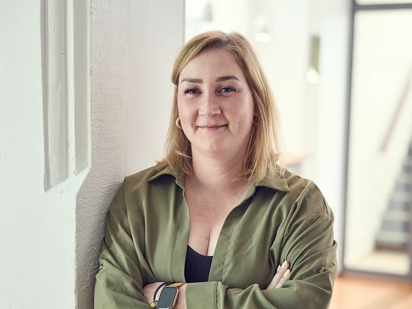 Eine Person mit blonden Haaren steht an einer Wand und lehnt mit verschränkten Armen. Sie trägt eine grüne Bluse und hat einen selbstbewussten Gesichtsausdruck. Im Hintergrund sind unscharfe Elemente eines modernen Innenraums erkennbar.