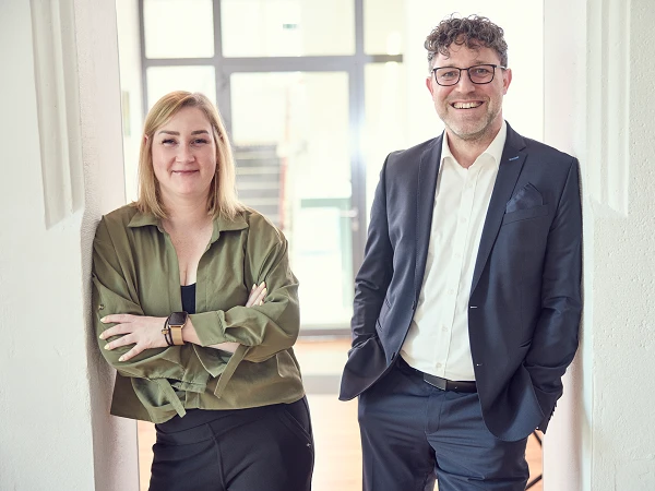 Zwei Personen stehen in einem hellen, modernen Raum; die Frau trägt eine grüne Bluse und der Mann einen Anzug, beide lächeln freundlich in die Kamera.