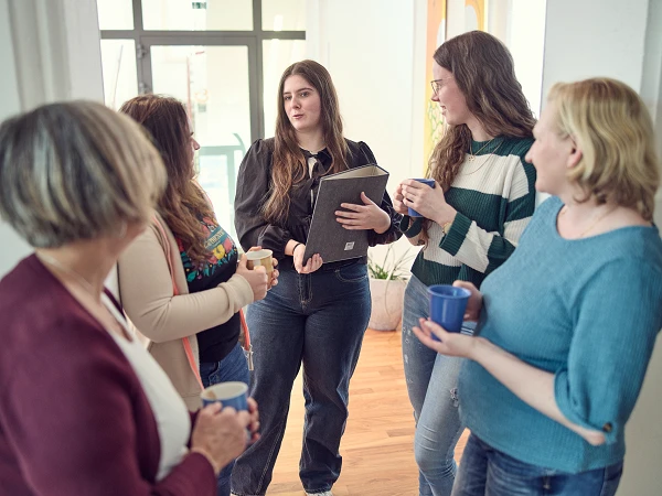 Sechs Frauen stehen in einem hellen Raum zusammen, während eine von ihnen ein Notizbuch hält und spricht. Die anderen halten Tassen in den Händen und hören aufmerksam zu. Pflanzen sind im Hintergrund sichtbar.