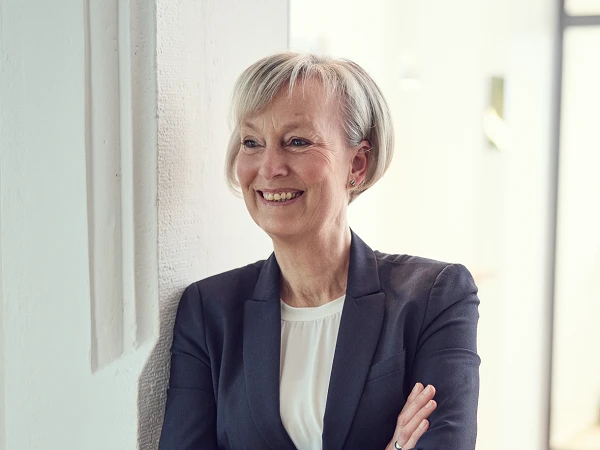 Eine lächelnde Frau mit kurzen, blonden Haaren steht an einer Wand, die Arme mühelos verschränkt, während sie in einem modernen Innenraum mit Holzfußboden und Treppe im Hintergrund positioniert ist. Sie trägt einen schwarzen Blazer über einem weißen Oberteil.