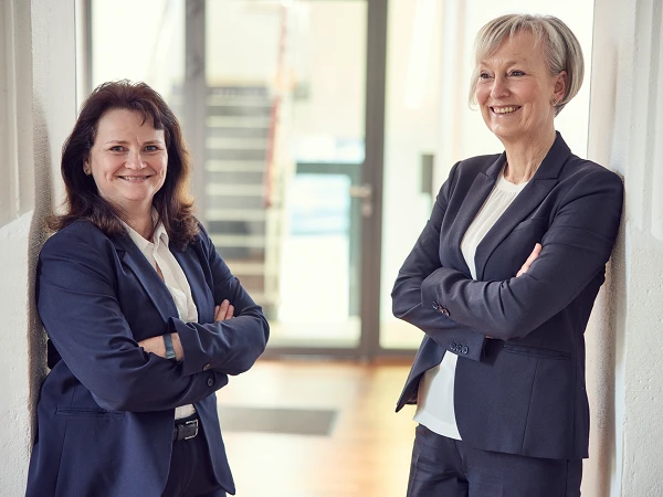 Zwei Frauen in formeller Kleidung stehen lächelnd nebeneinander in einem modernen Büroflur. Sie haben die Arme verschränkt und wirken freundlich und selbstbewusst. Im Hintergrund sind Treppen und ein Fenster zu sehen.