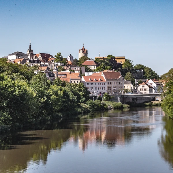 Eine malerische Aussicht auf eine Stadt, die am Ufer eines Flusses liegt, umgeben von grünen Bäumen und Hügeln, mit historischen Gebäuden und Kirchen im Hintergrund. Der Himmel ist klar und blau, während sich die Stadt im Wasser spiegelt.