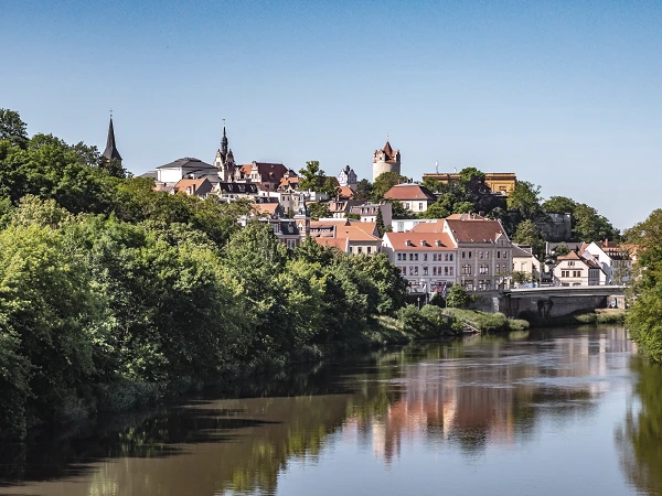 Eine malerische Aussicht auf eine Stadt, die am Ufer eines Flusses liegt, umgeben von grünen Bäumen und Hügeln, mit historischen Gebäuden und Kirchen im Hintergrund. Der Himmel ist klar und blau, während sich die Stadt im Wasser spiegelt.