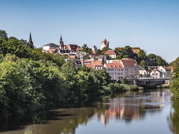 Eine malerische Stadtansicht mit bunt gedeckten Häusern und einem historischen Turm, umgeben von üppigem Grün und reflektiert im ruhigen Wasser eines Flusses.