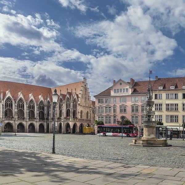 Der Marktplatz zeigt historische Gebäude mit einem gotischen Rathaus links, bunten Fassaden rechterhand und einer zentralen Brunnenanlage. Der Himmel ist mit einigen Wolken bedeckt, und ein Bus steht am Rand der Fläche.