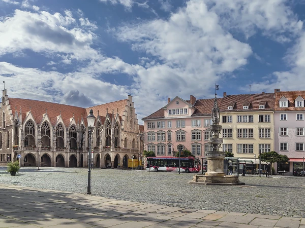 Der Marktplatz zeigt historische Gebäude mit einem gotischen Rathaus links, bunten Fassaden rechterhand und einer zentralen Brunnenanlage. Der Himmel ist mit einigen Wolken bedeckt, und ein Bus steht am Rand der Fläche.