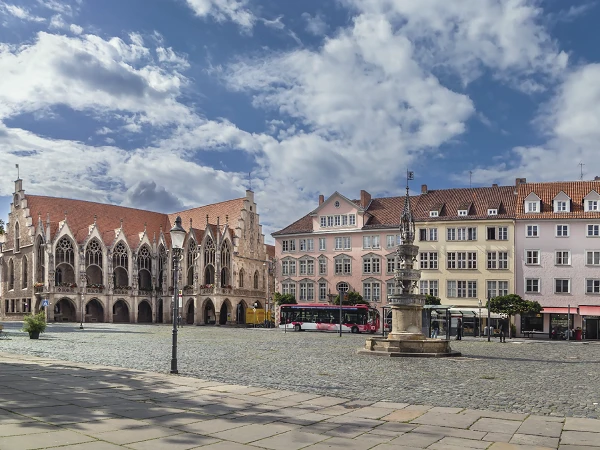 Blick auf einen historischen Platz mit gotischen Gebäuden, darunter ein Rathaus mit spitzbogen Fenstern, einer neugotischen Statue in der Mitte und pastelrosa Häusern im Hintergrund, unter einem klaren, bewölkten Himmel.