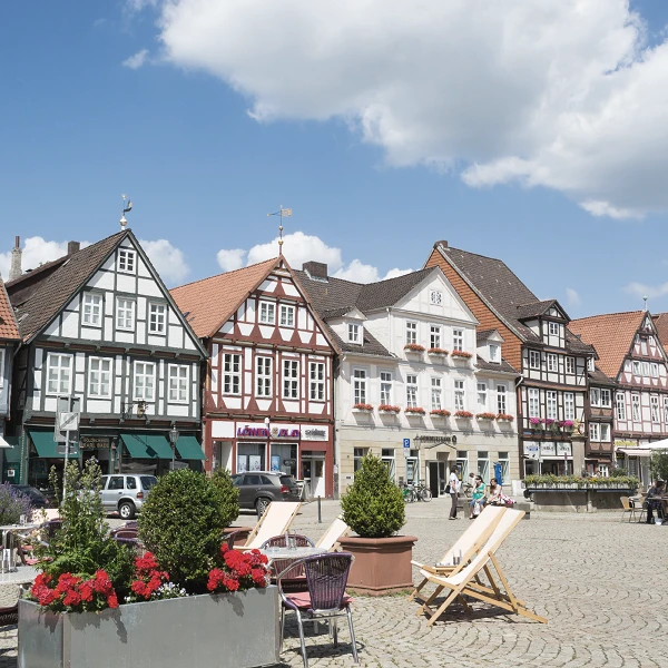 Ein malerischer Platz mit historischen Fachwerkhäusern, die in verschiedenen Farben gestaltet sind. Im Vordergrund befinden sich gemütliche Liegestühle und Blumenbeete mit bunten Blumen. Der Himmel ist blau mit einigen Wolken.