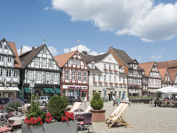 Ein malerischer Platz mit historischen Fachwerkhäusern, die in verschiedenen Farben gestaltet sind. Im Vordergrund befinden sich gemütliche Liegestühle und Blumenbeete mit bunten Blumen. Der Himmel ist blau mit einigen Wolken.