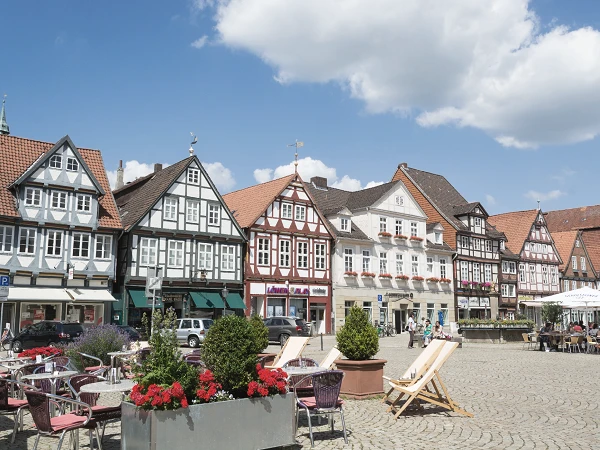Der historische Marktplatz zeigt eine Reihe von bunten, zum Teil mit Fachwerk versehenen Gebäuden und gemütlichen Sitzgelegenheiten im Freien, umgeben von blühenden Pflanzen und einem klaren blauen Himmel.