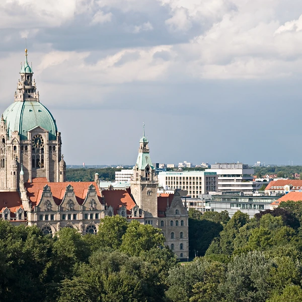 Die Stadtsilhouette von Hannover zeigt im Vordergrund das historische Rathaus mit einer markanten Kuppel, während im Hintergrund moderne Gebäude und eine bewaldete Landschaft zu sehen sind. Der Himmel ist bewölkt und gibt der Szene einen dramatischen Anstrich.