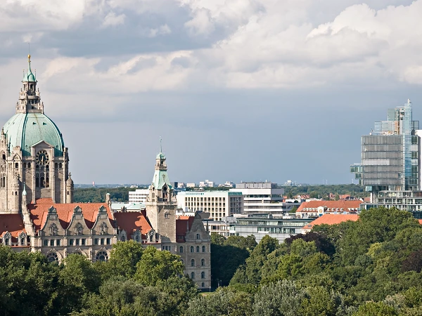 Die Stadtsilhouette von Hannover zeigt im Vordergrund das historische Rathaus mit einer markanten Kuppel, während im Hintergrund moderne Gebäude und eine bewaldete Landschaft zu sehen sind. Der Himmel ist bewölkt und gibt der Szene einen dramatischen Anstrich.