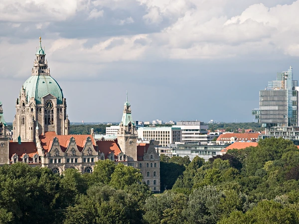Die Skyline von Hannover mit dem historischen Rathaus im Vordergrund und modernen Bürogebäuden im Hintergrund, umgeben von Bäumen und einem bewölkten Himmel.