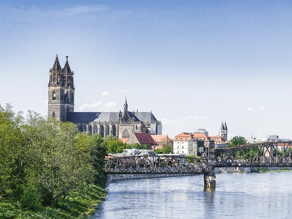 Die imposante Kirche mit einem hohen Turm überblickt eine Stadtlandschaft, während eine Eisenbahnbrücke über den Fluss führt und umliegendes Grün die Szenerie umrahmt.