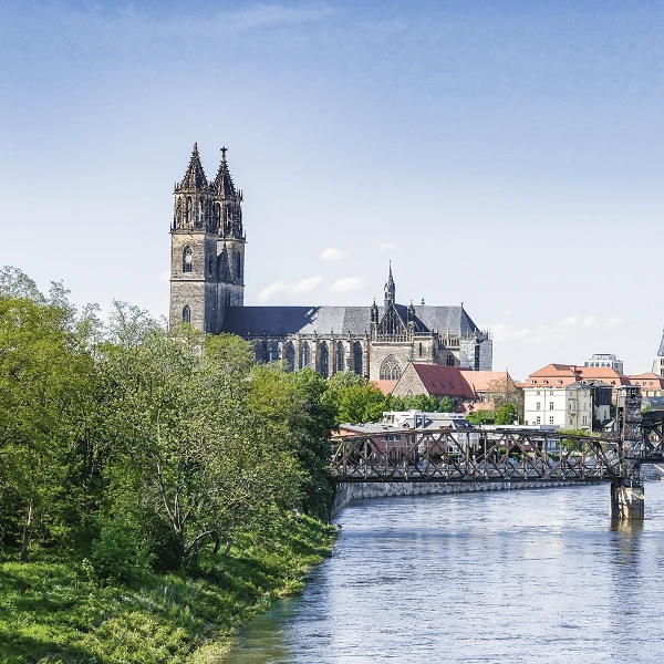 Die Skyline einer Stadt mit einer markanten Kirche im Hintergrund, die von grünen Bäumen und einem Fluss umgeben ist, sowie einer Brücke, die über das Wasser führt.