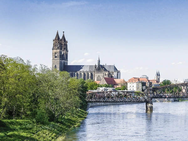 Die Skyline einer Stadt mit einer markanten Kirche im Hintergrund, die von grünen Bäumen und einem Fluss umgeben ist, sowie einer Brücke, die über das Wasser führt.