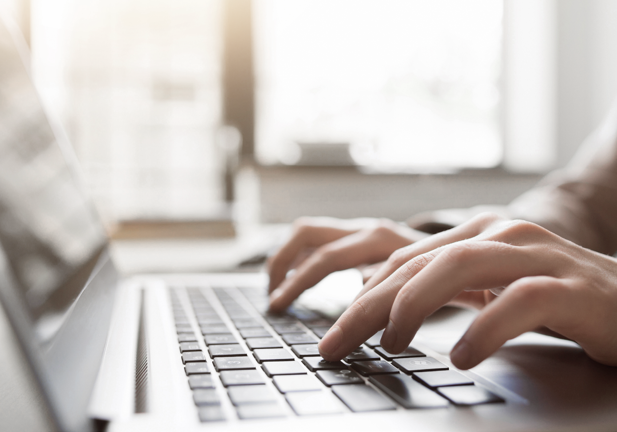 Eine Nahaufnahme von Händen, die auf einer Laptop-Tastatur tippen, während im Hintergrund weiches, natürliches Licht durch ein Fenster strömt.