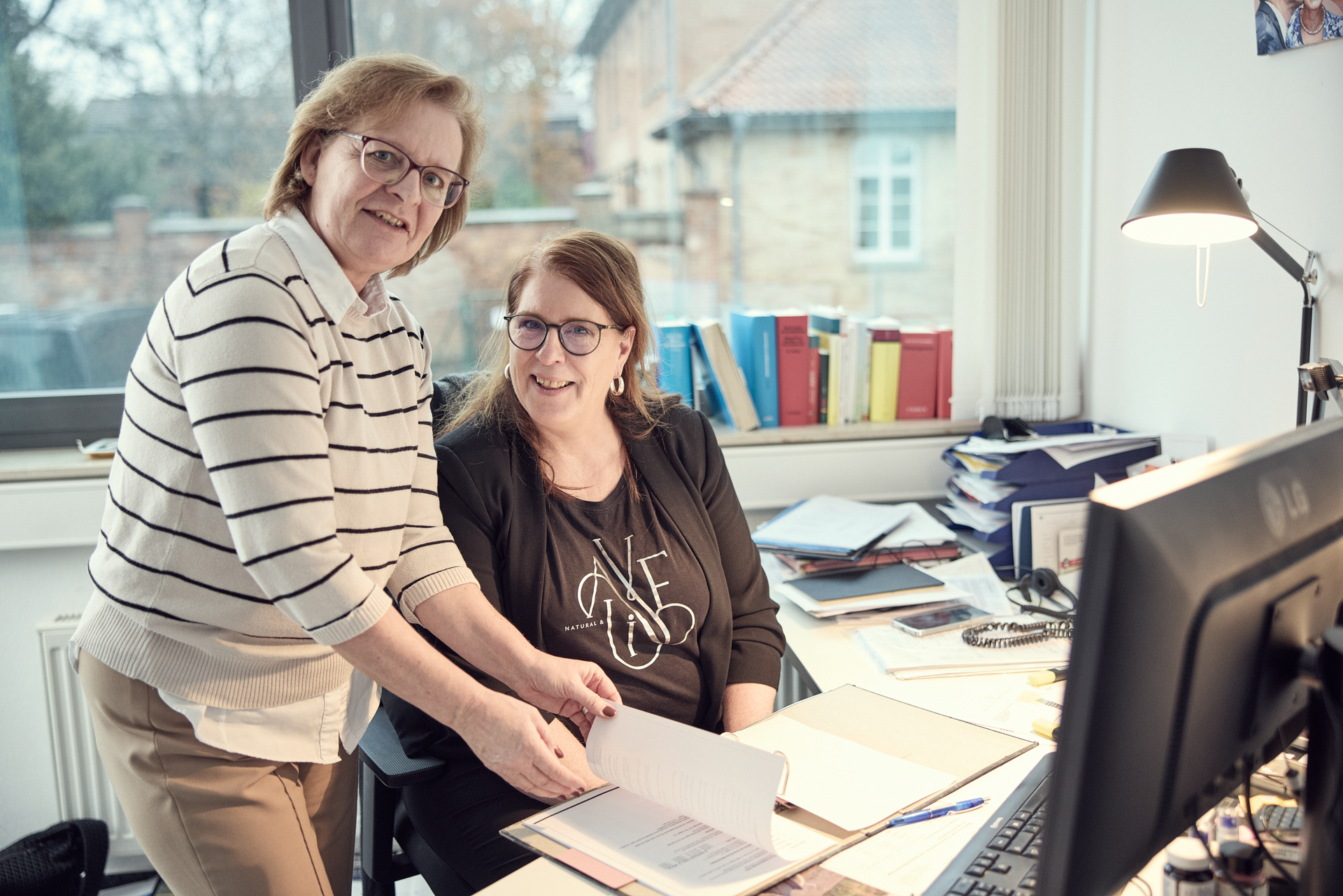 Zwei Frauen stehen und sitzen an einem Schreibtisch in einem Büro, während sie in ein Dokument schauen. Im Hintergrund sind Bücherregale und eine große Fensterfront sichtbar.
