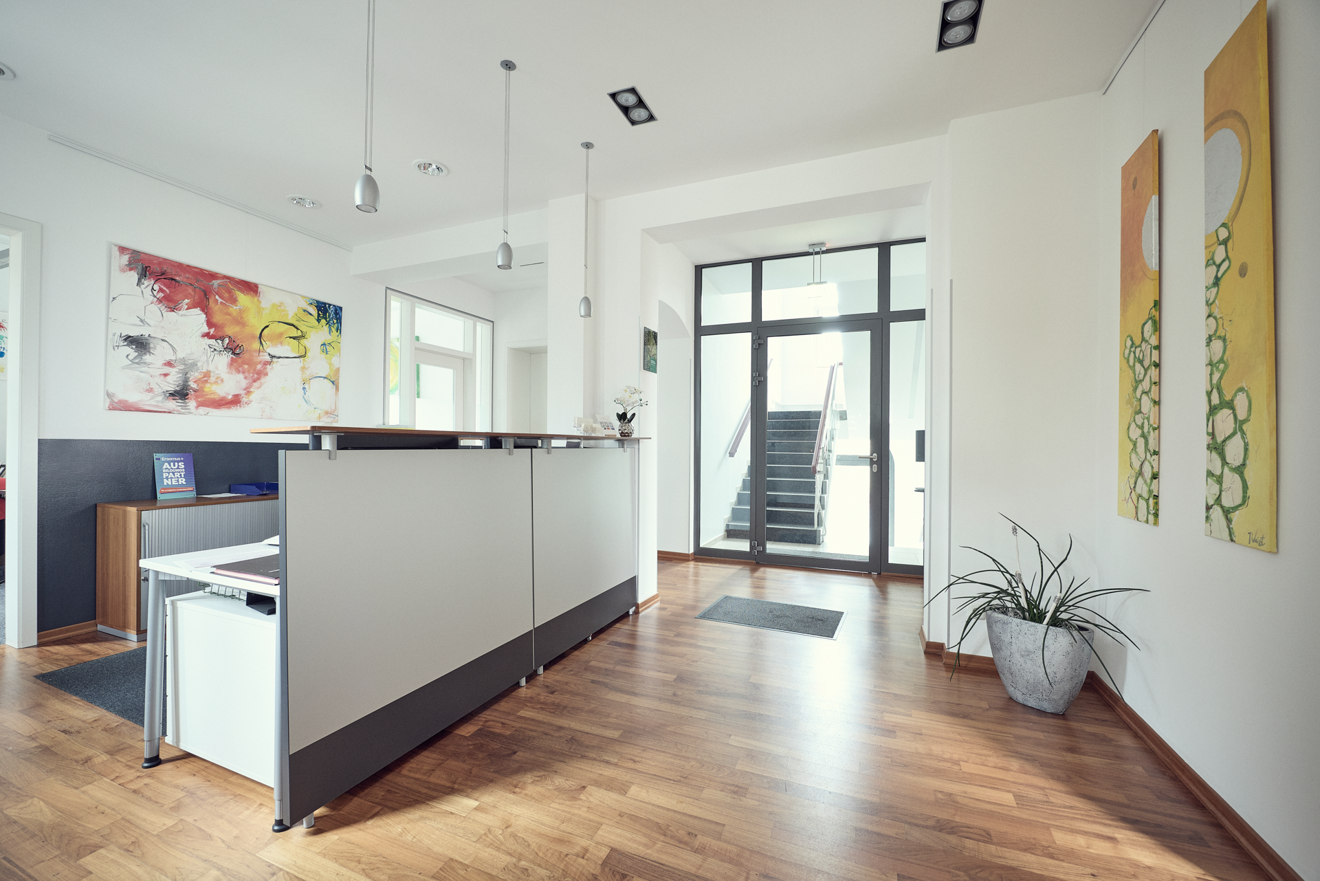Der Empfangsbereich eines modernen Büros mit einem hellen, offenen Raumdesign. Eine Empfangstheke steht im Vordergrund, und im Hintergrund sind zwei farbenfrohe Gemälde sowie eine Treppe sichtbar, die zu einem weiteren Bereich führt. Eine Pflanze steht in der Ecke, und es gibt große Fenster, die viel Tageslicht hereinlassen.