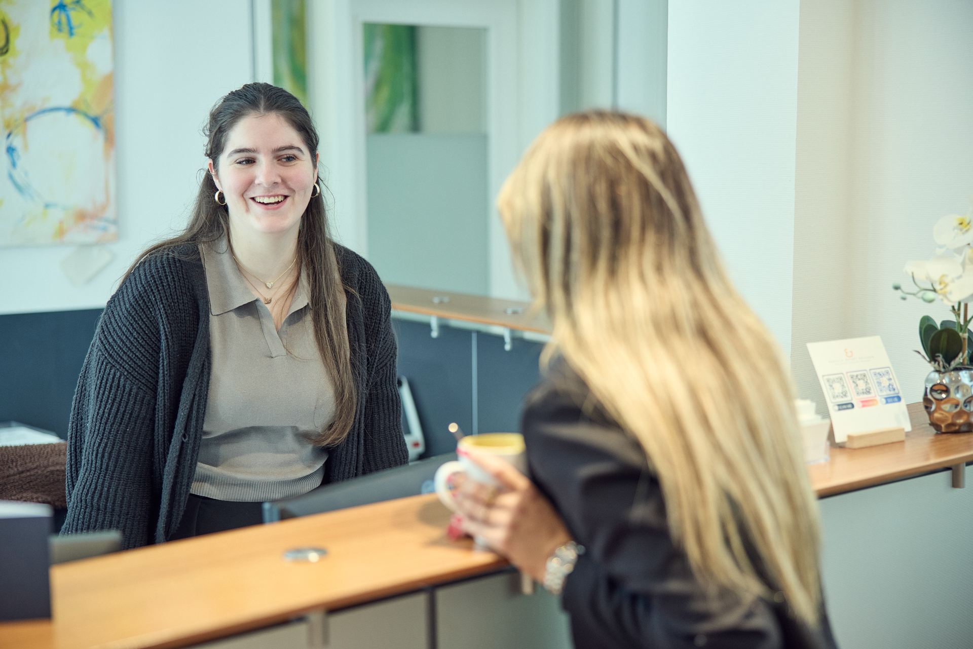 Eine junge Frau mit langen, braunen Haaren und einem lächelnden Gesicht steht an einem Empfangstresen und spricht freundlich mit einer anderen Person, die mit dem Rücken zur Kamera steht. Im Hintergrund sind einige Dekorationen und ein paar Informationsmaterialien sichtbar.