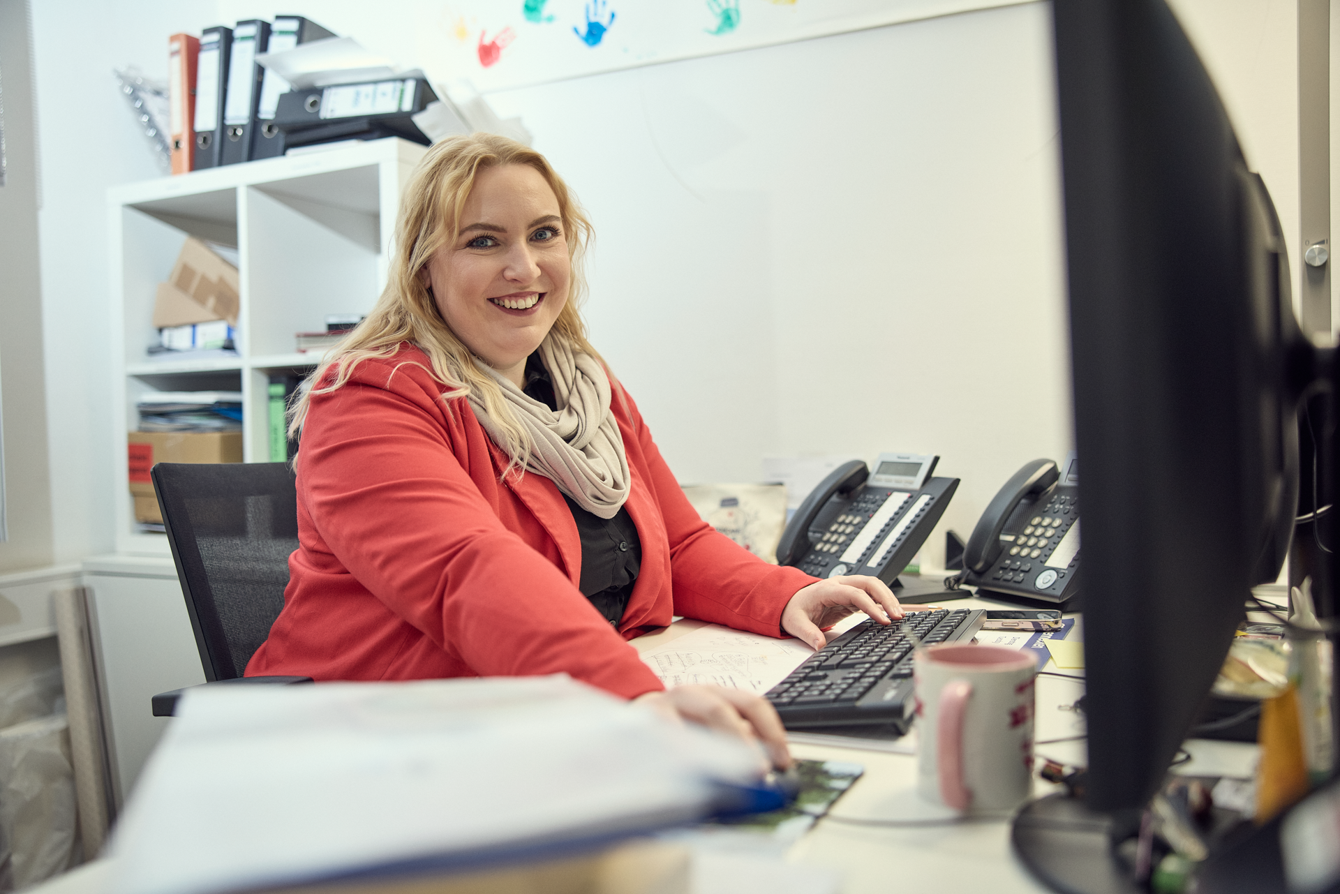 Eine lächelnde Frau in einem roten Blazer sitzt an einem Schreibtisch, tippt auf einer Tastatur und schaut in die Kamera. Im Hintergrund sind mehrere Telefone und Gegenstände auf dem Schreibtisch sichtbar.