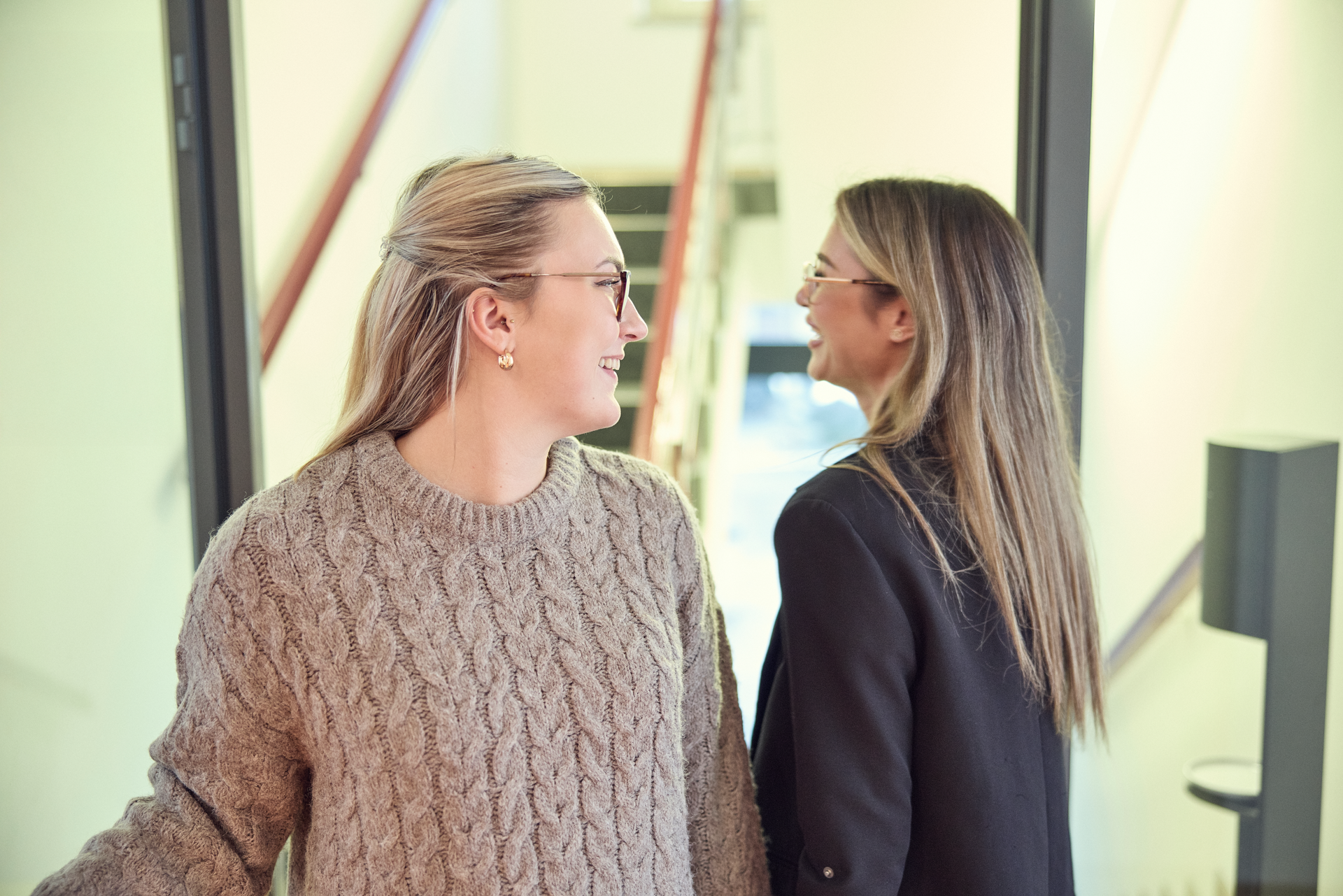 Zwei Frauen stehen in einem hellen Raum und blicken freundlich zueinander. Eine Frau trägt einen großen, strickartigen Pullover, während die andere eine dunkle Jacke hat. Im Hintergrund sind Treppen und eine moderne Architektur sichtbar.