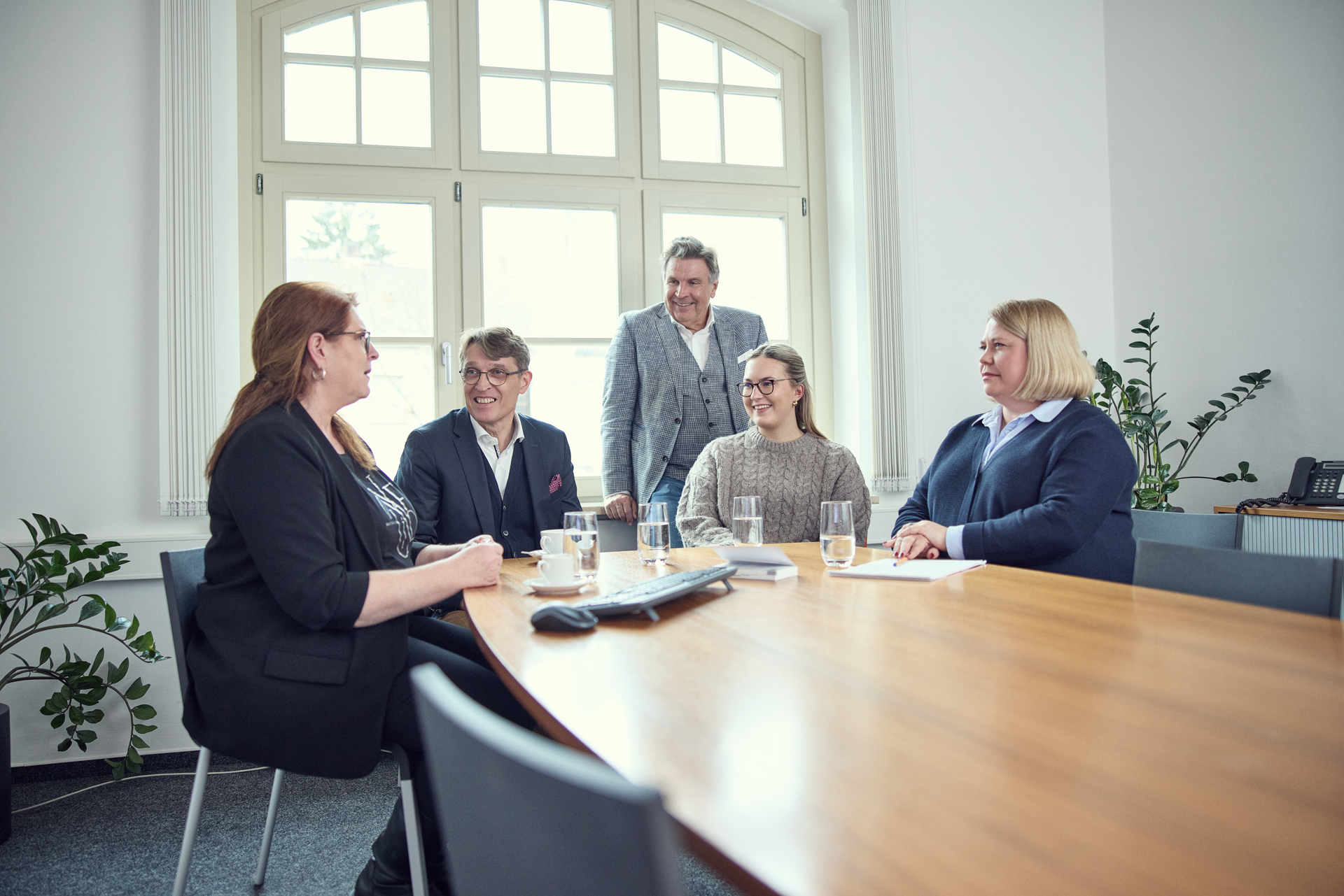 Eine Gruppe von fünf Personen sitzt an einem Konferenztisch in einem hellen Raum mit großen Fenstern. Vier Personen sind am Tisch platziert und in ein Gespräch vertieft, während eine Person hinter ihnen steht und aufmerksam lauscht. Auf dem Tisch stehen Gläser mit Wasser und Unterlagen. Pflanzen sind im Hintergrund zu sehen.