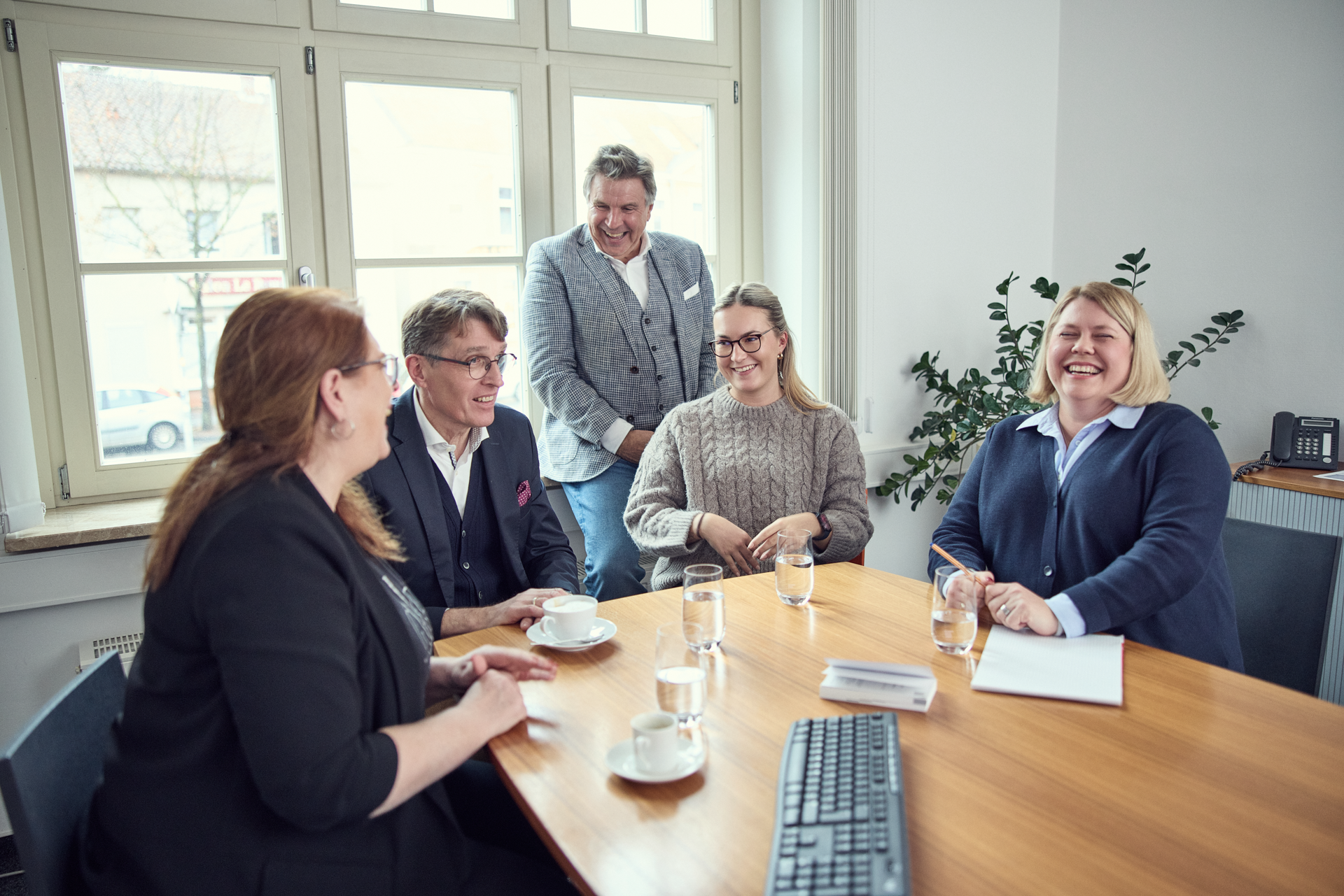 Eine Gruppe von fünf Personen sitzt an einem Tisch in einem hellen Raum, während sie sich unterhalten und Lächeln austauschen. Auf dem Tisch stehen Kaffeetassen und Gläser mit Wasser. Jeder ist in formeller Kleidung, und im Hintergrund sind Fenster und eine Pflanze zu sehen.