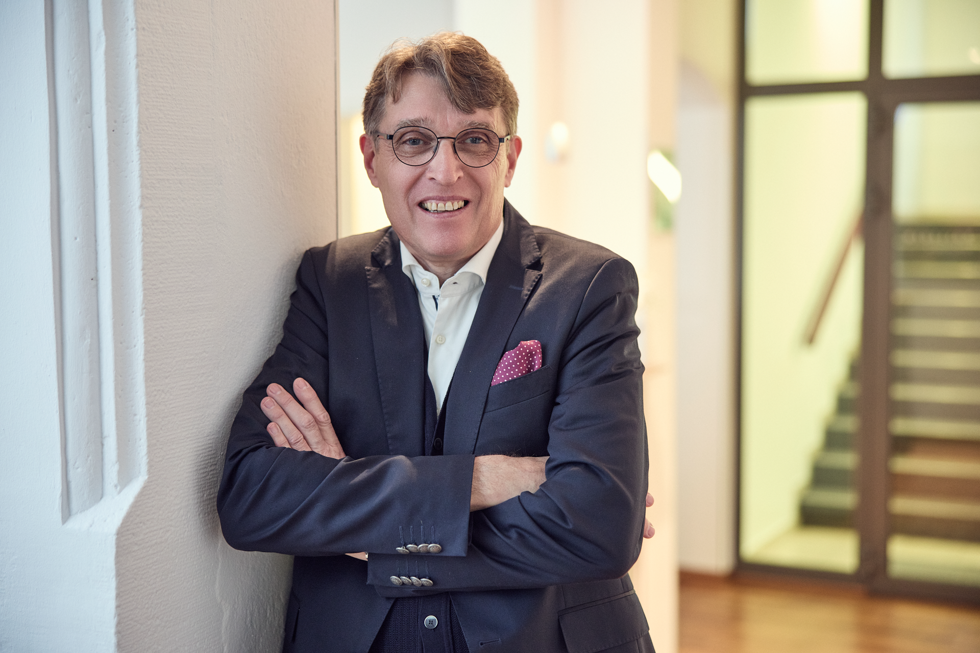 Ein Mann mit Brille und grauen Haaren leans an einer Wand in einem modernen Innenraum und lächelt. Er trägt einen eleganten Anzug mit einer Weste und hat ein rotes Taschentuch in der Brusttasche. Im Hintergrund sind Treppen und ein heller Flur sichtbar.
