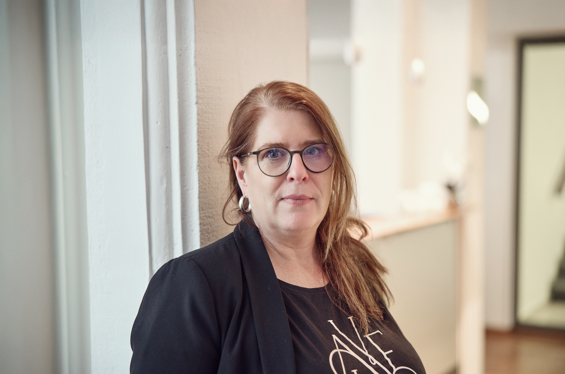 Eine Frau mit langen, braunen Haaren und Brille steht an einer Wand in einem modernen Innenraum und schaut direkt in die Kamera. Sie trägt ein schwarzes Oberteil mit einem weißen Schriftzug und eine schwarze Jacke. Im Hintergrund sind unscharfe Möbel und Lichtquellen zu erkennen.