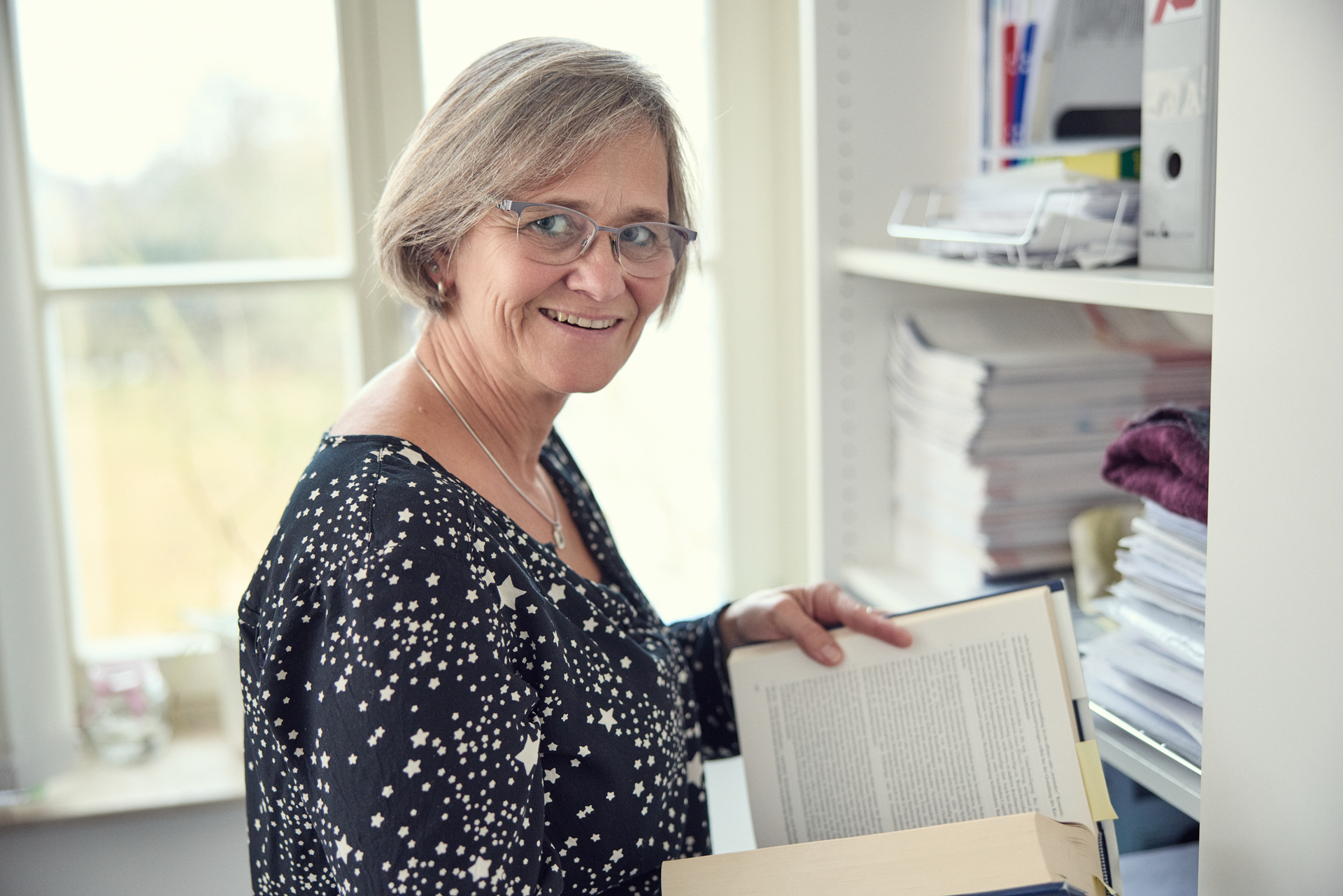 Eine lächelnde Frau mit Brille steht neben einem Regal und hält ein aufgeschlagenes Buch in der Hand, während sie nach einem weiteren Buch greift. Im Hintergrund sind Ordner und Papierstapel zu sehen.
