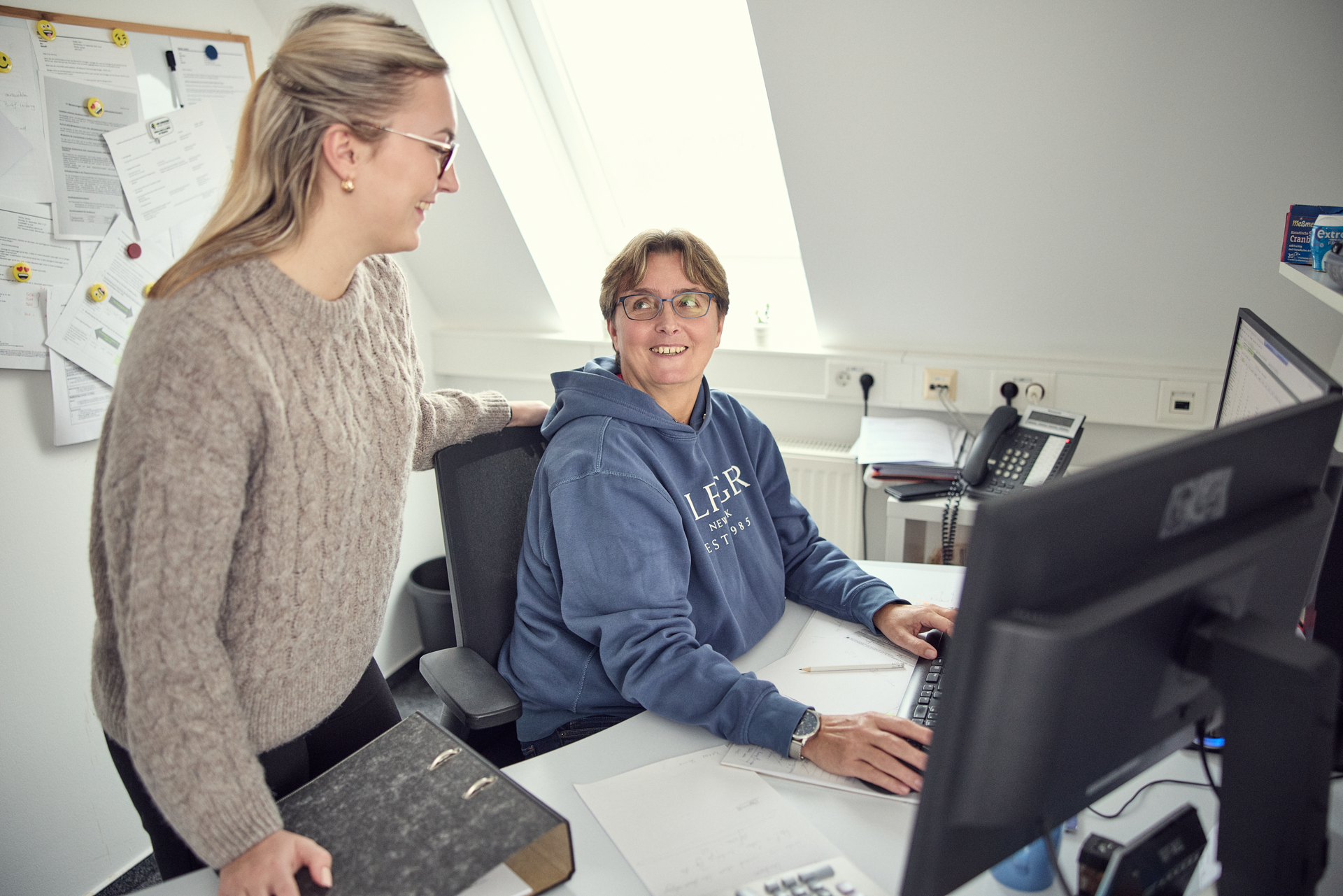 Zwei Frauen in einem Büro kommunizieren, während eine von ihnen am Computer sitzt und die andere hinter ihr steht und lächelt. Die sitzende Frau trägt einen blauen Kapuzenpullover, während die stehende Frau einen beigen Pullover trägt und eine Brille auf hat. Ein Schreibtisch mit Dokumenten und einem Computer ist im Vordergrund sichtbar.