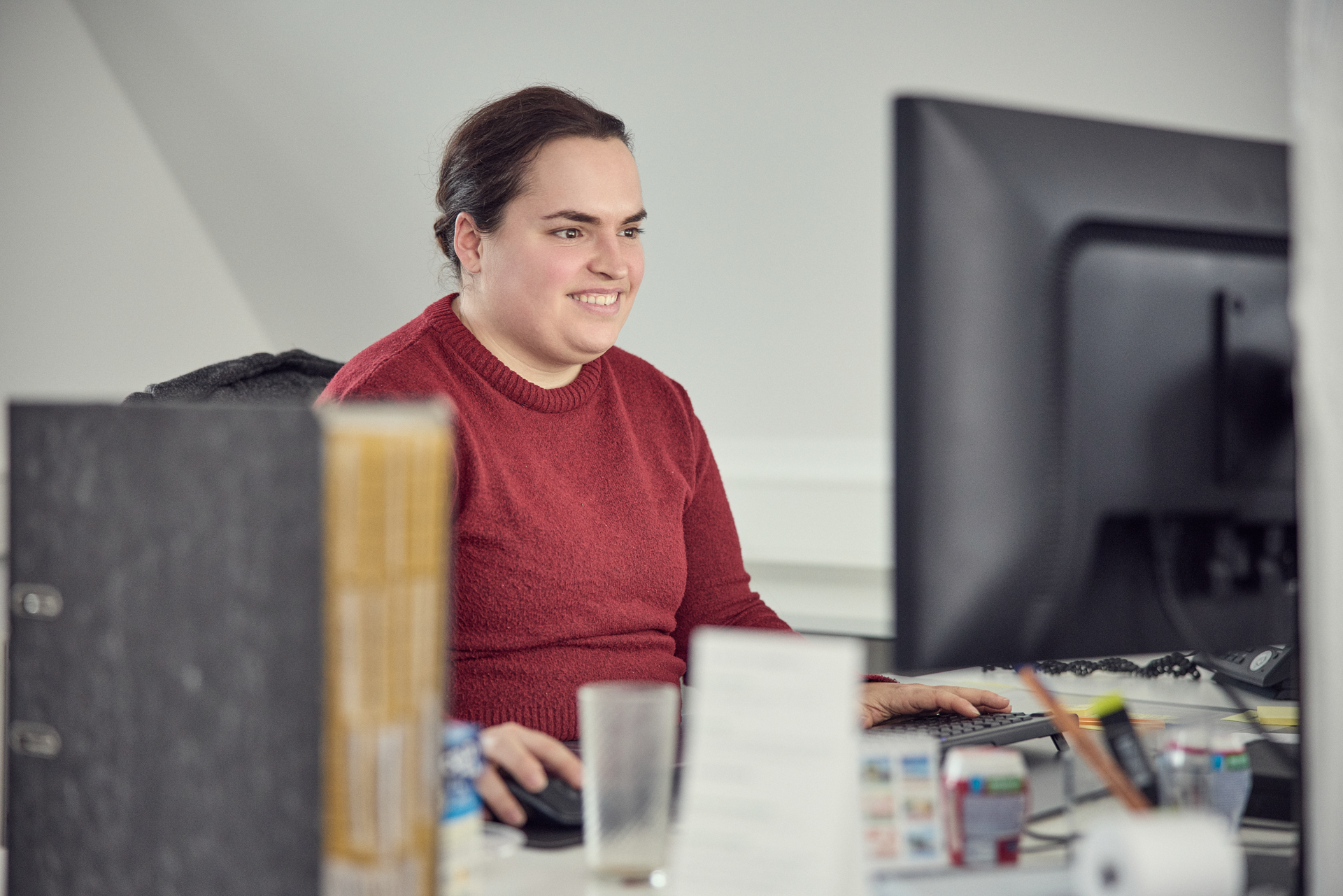 Eine Person mit langen, dunklen Haaren und einem roten Pullover sitzt an einem Schreibtisch und arbeitet konzentriert an einem Computer. Auf dem Tisch befinden sich verschiedene Büromaterialien und ein leeres Glas.