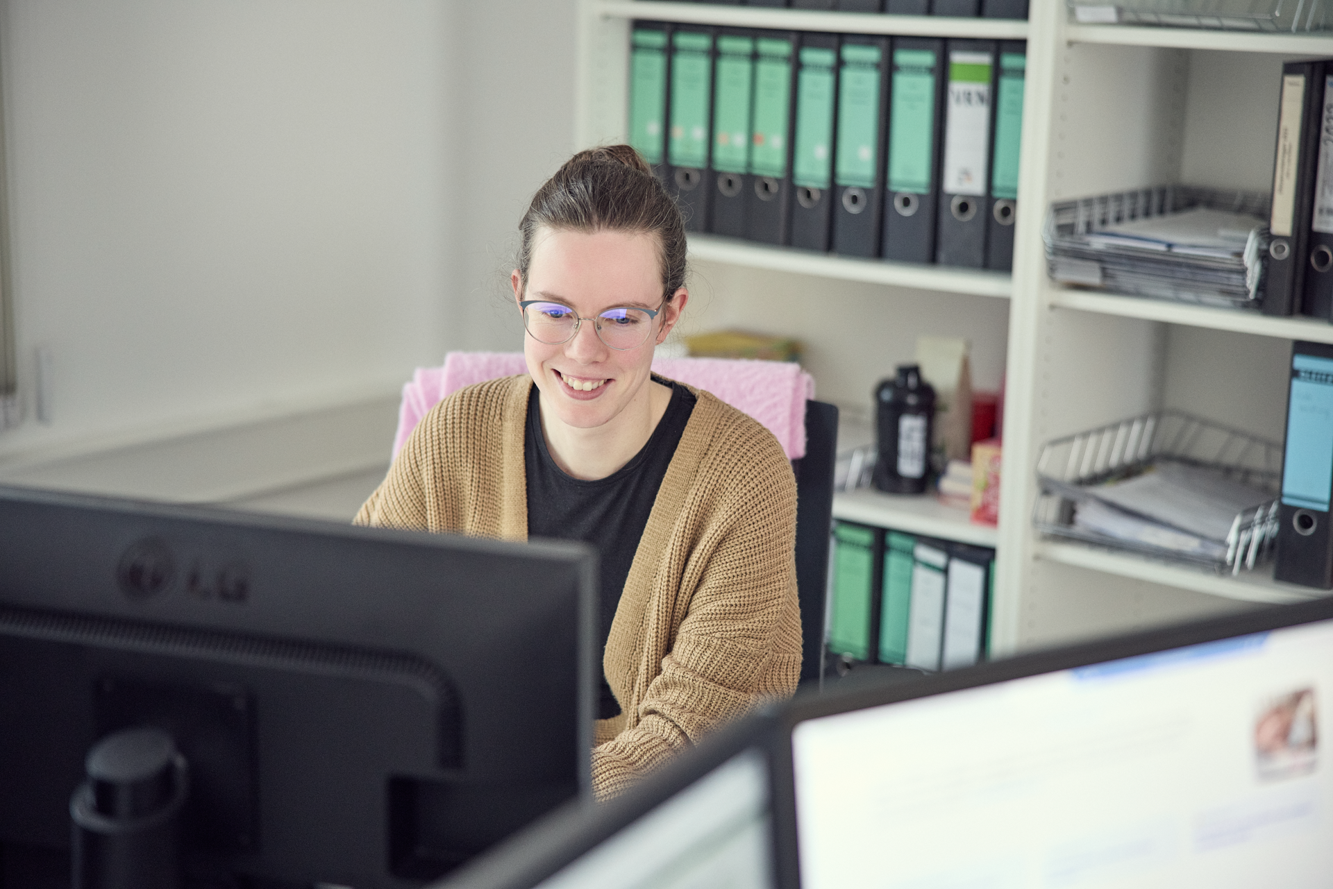 Eine junge Frau mit Brille sitzt an einem Schreibtisch und lächelt, während sie auf einen Computerbildschirm schaut. Im Hintergrund sind Aktenordner und Büromaterial zu sehen.
